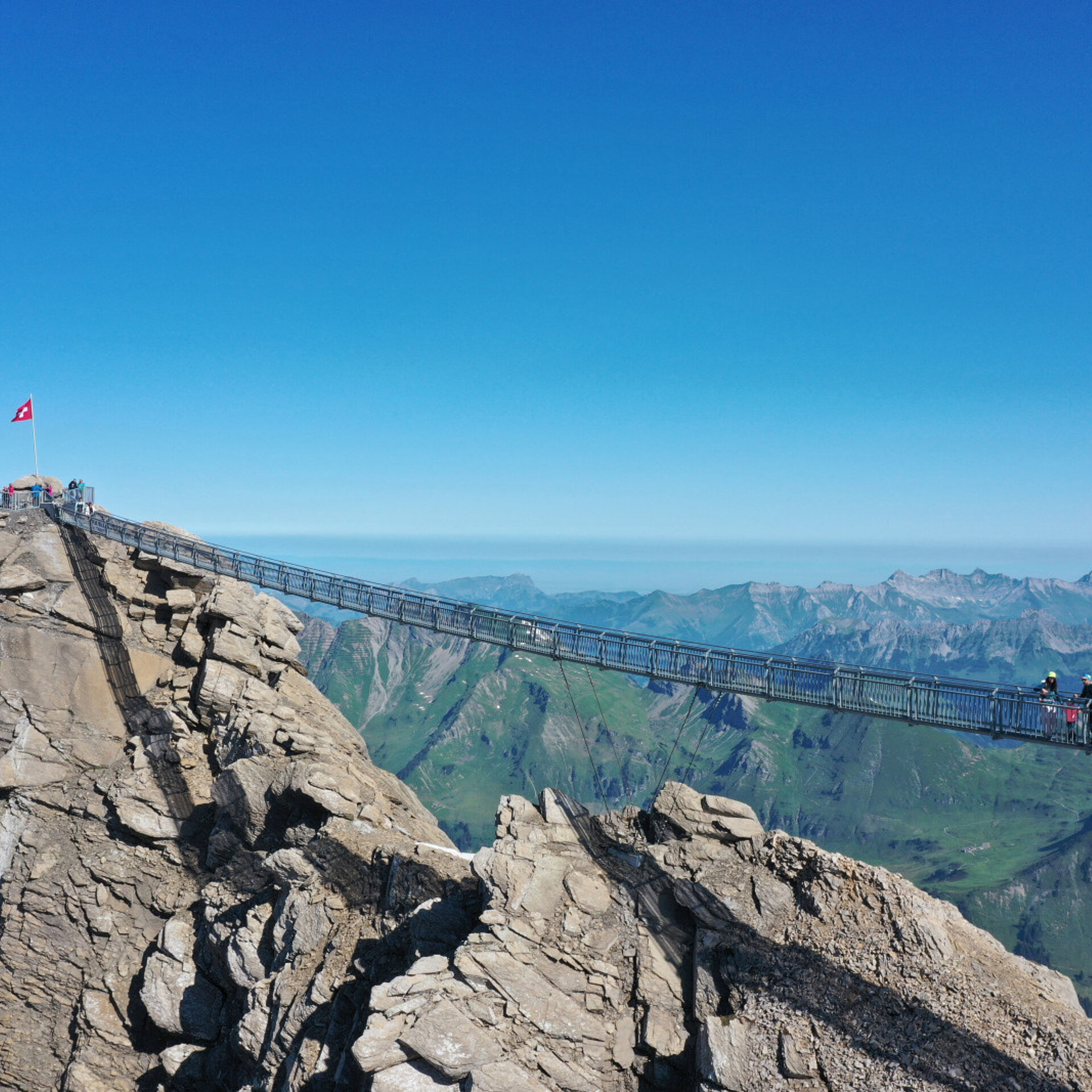 Eine Hängebrücke, welche zwei felsige Gipfel verbindet. Im Hintergrund ein sommerliches Bergpanorama.