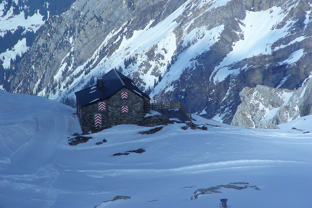 Grau gemauerte Berghütte mit schwarzem Dach und geschlossenen rot-weiss gestreiften Fensterläden und einer Terasse in winterlicher Berglandschaft.