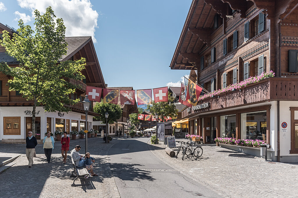 Promenade mit blumengeschmückten Chalets, Fussgängern, einem Mann und einem Kind auf einer Sitzbank, Bäume und Strassenlaternen, in der Mitte hängen Fahnen von der Schweiz, dem Kanton Bern und der Gemeinde Saanen.