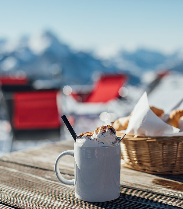 Auf einem Tisch steht eine heisse Schokolade mit Schlagrahm und ein Körbchen mit Pommes. Dahinter sieht man verschwommen ein verschneites Bergpanorama.