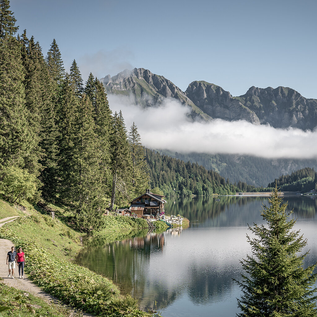 Zwei Wandrer:innen auf einem Fussweg entlang eines Bergsees, im Hintergrund Berge mit einer Nebelbank davor.