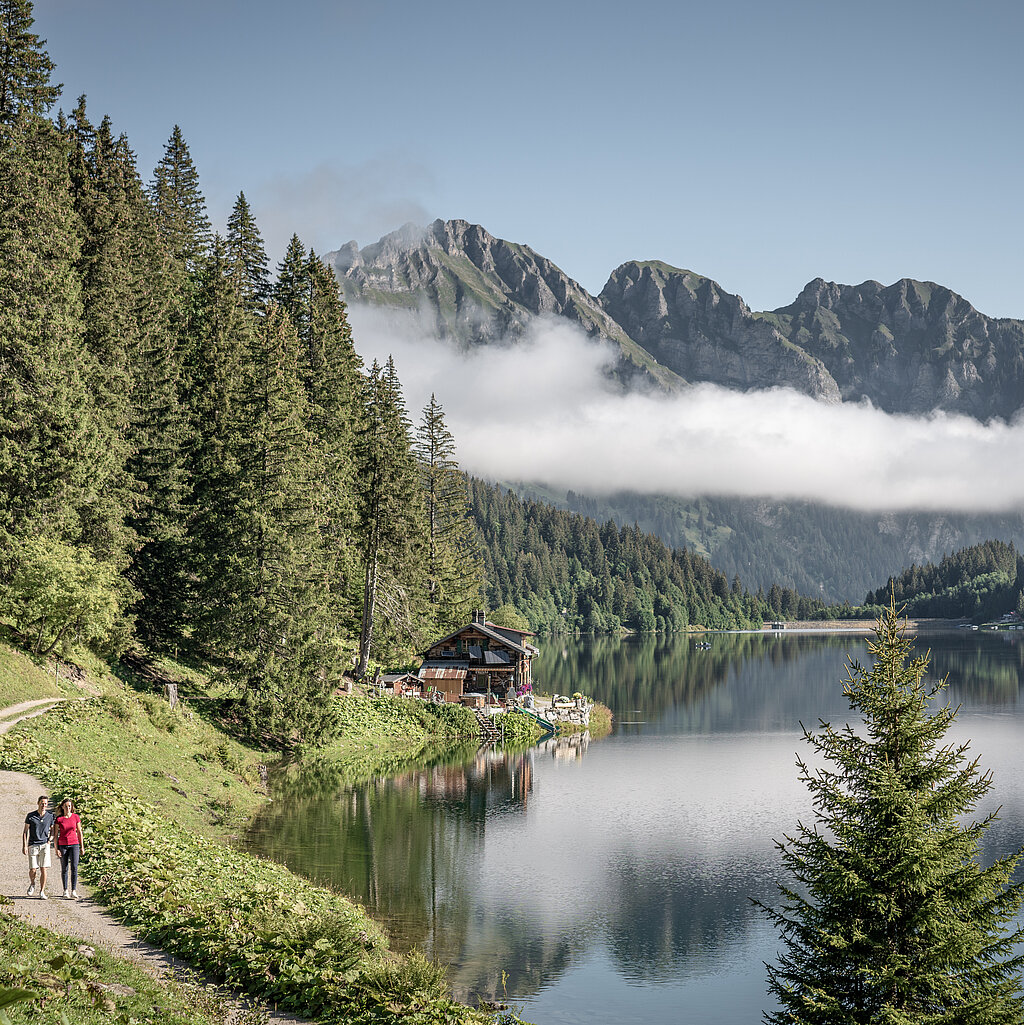 Zwei Wandrer:innen auf einem Fussweg entlang eines Bergsees, im Hintergrund Berge mit einer Nebelbank davor.