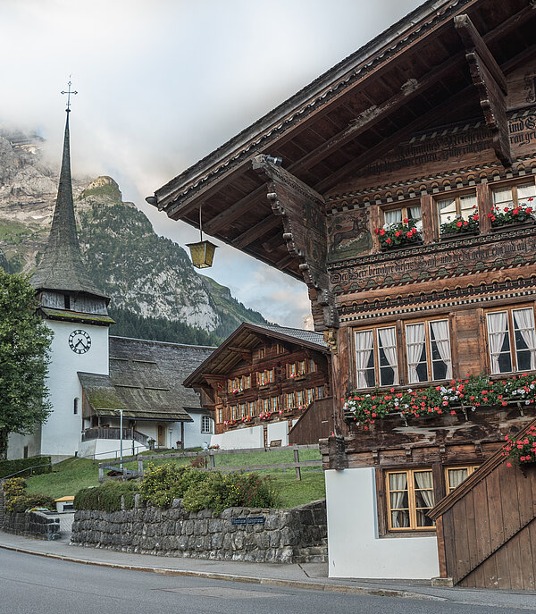 Im Vordergrund sieht man das Hotel Bären in Gsteig. Dies ist ein sehr altes Holzchalet mit roten Geranien vor den Fenstern und alten Zeichnungen an der Front. Im Hintergrund ist die Kirche Gsteig sichtbar.